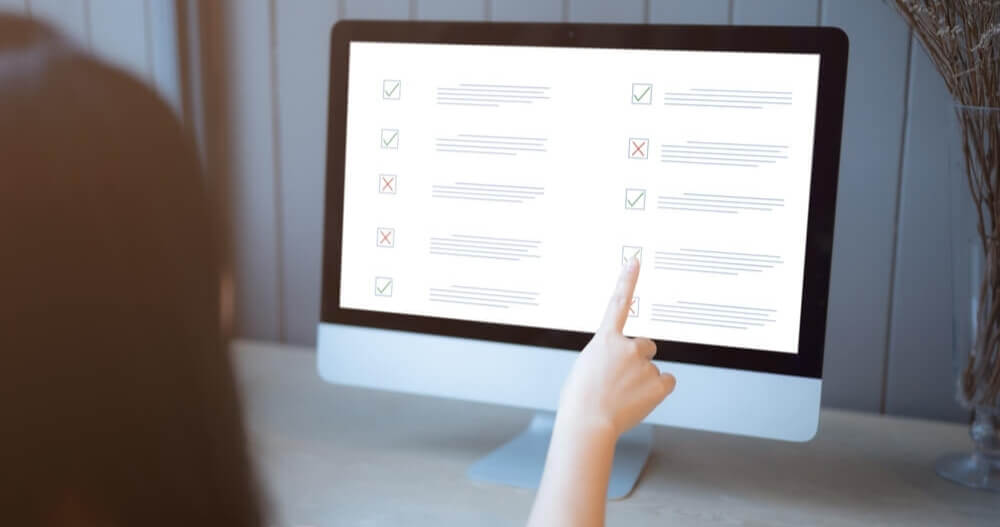 A person pointing at a computer screen displaying a checklist with green checkmarks and red crosses, suggesting task completion or assessment review.